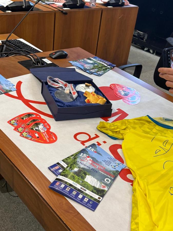 A conference table displaying a navy box containing a medal and ribbons, a yellow athletic shirt, brochures about a marathon event, and promotional stickers, with a background of wooden cabinets and office equipment.