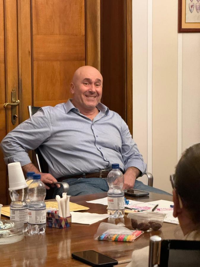 A smiling man with a bald head sitting in a chair at a meeting table, surrounded by items like water bottles, paper, and colorful snacks.