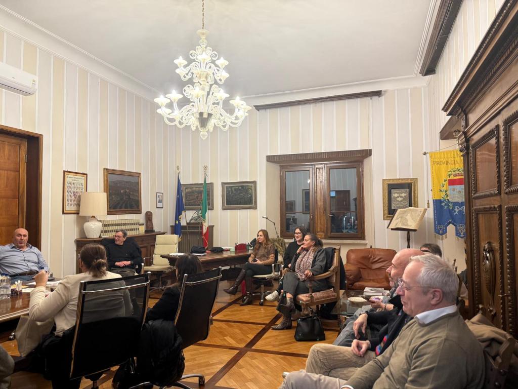 A meeting taking place in a well-decorated room with a chandelier, featuring several people seated around a large table, engaged in conversation. The room includes artwork, flags, and wooden furniture.