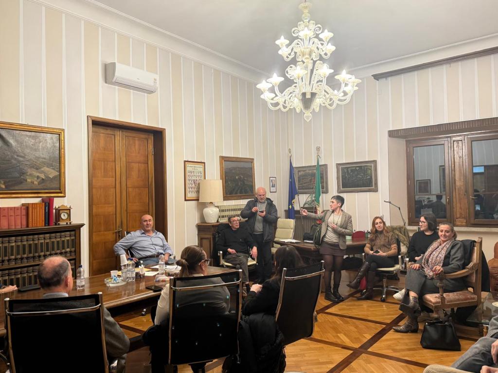 A group of people gathered in a formal meeting room, engaged in discussion. The room features wooden furnishings, a large table, and decorative paintings on the walls.