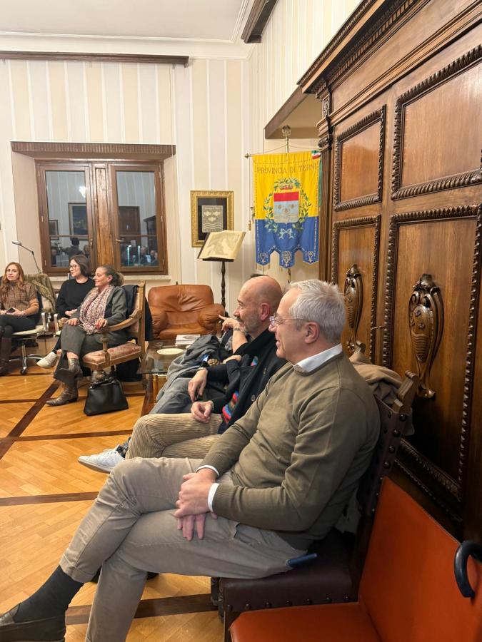 A group of people sitting in a well-decorated room with wooden furnishings and a banner hanging on the wall. Some attendees are engaged in conversation while others are listening.