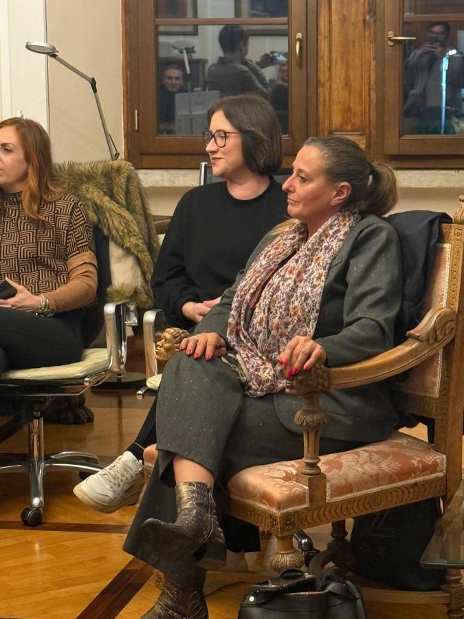 A group of three women sitting in a cozy indoor setting, with wooden windows in the background. One woman is wearing glasses and a black sweater, while another is in a patterned top. The third woman, sitting in an ornate chair, is dressed in gray and has a patterned scarf.