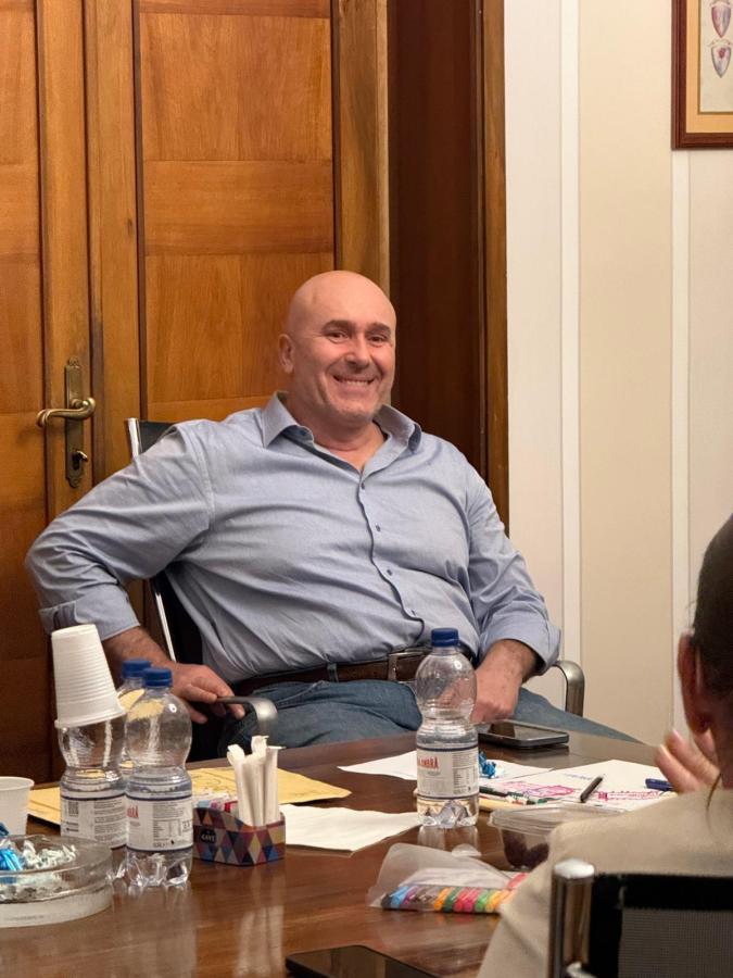 A smiling bald man sitting at a wooden table surrounded by water bottles, papers, and colorful party favors in a well-lit room.