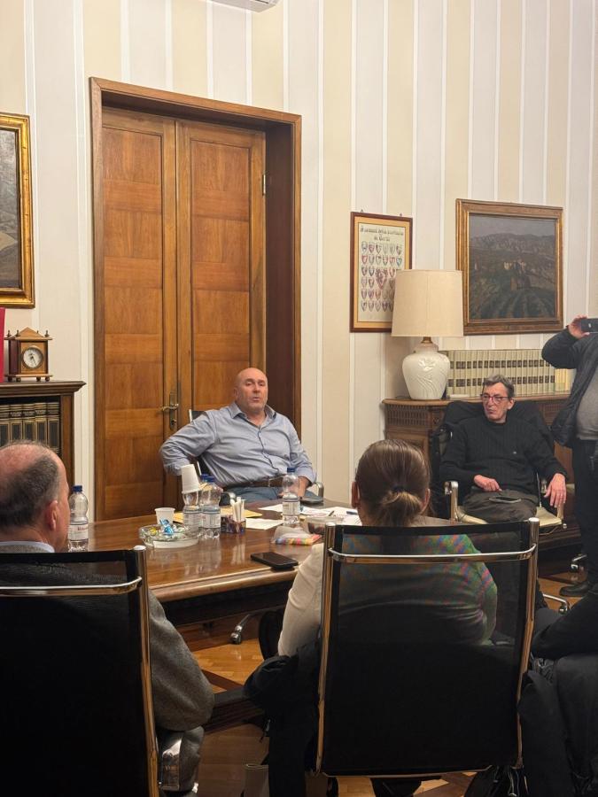 A group of people seated around a conference table in a well-decorated room, engaged in discussion. One person is speaking while others listen attentively.