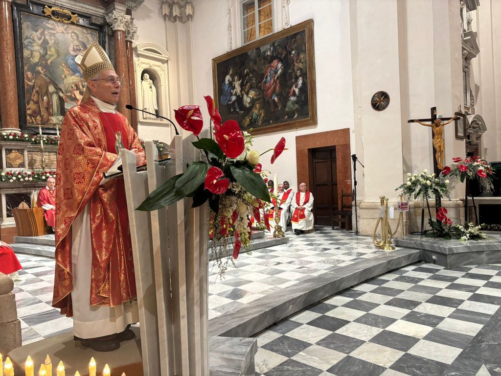 A priest in ornate vestments delivers a sermon at the altar of a church, with floral decorations and seated clergy members in the background.