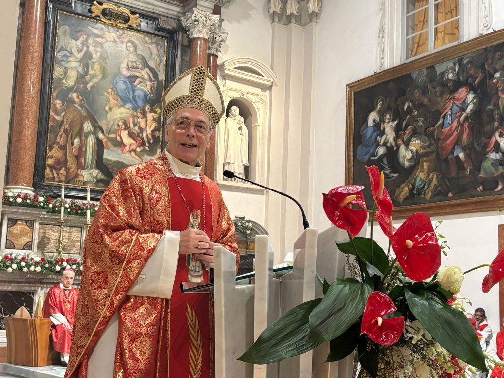 A clergy member in elaborate red robes and a mitre speaks at a lectern in a church, with vibrant floral arrangements in the foreground and religious paintings in the background.