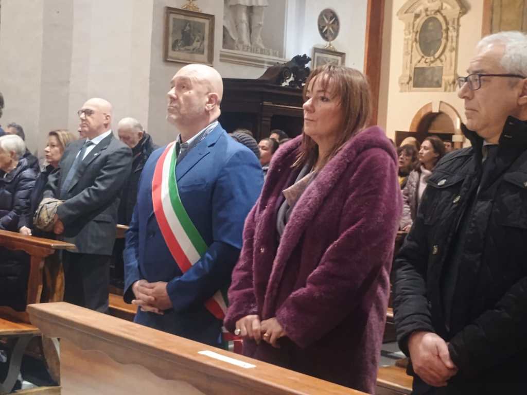 A group of people standing in a church during a ceremony, with a man wearing a suit and a tricolor sash in the foreground.