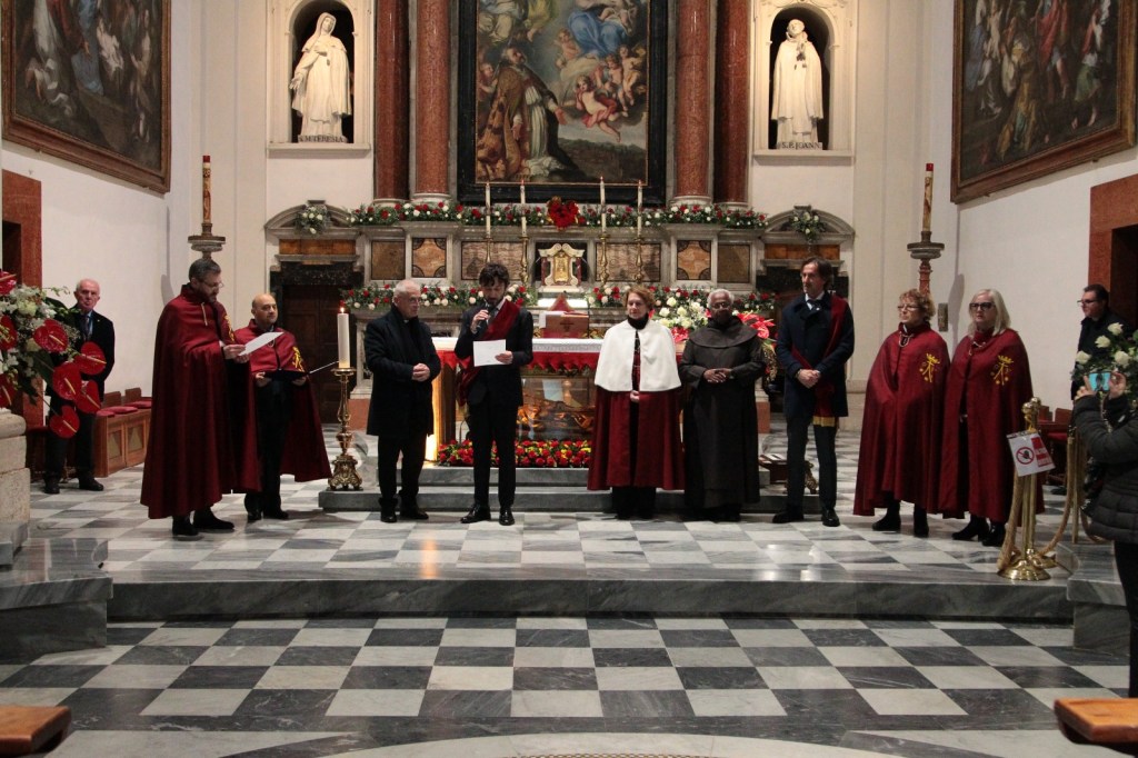 A group of people in ceremonial attire standing in a church, with an altar decorated with flowers. Some individuals are holding papers, while others observe. The interior features religious artwork and a statue.
