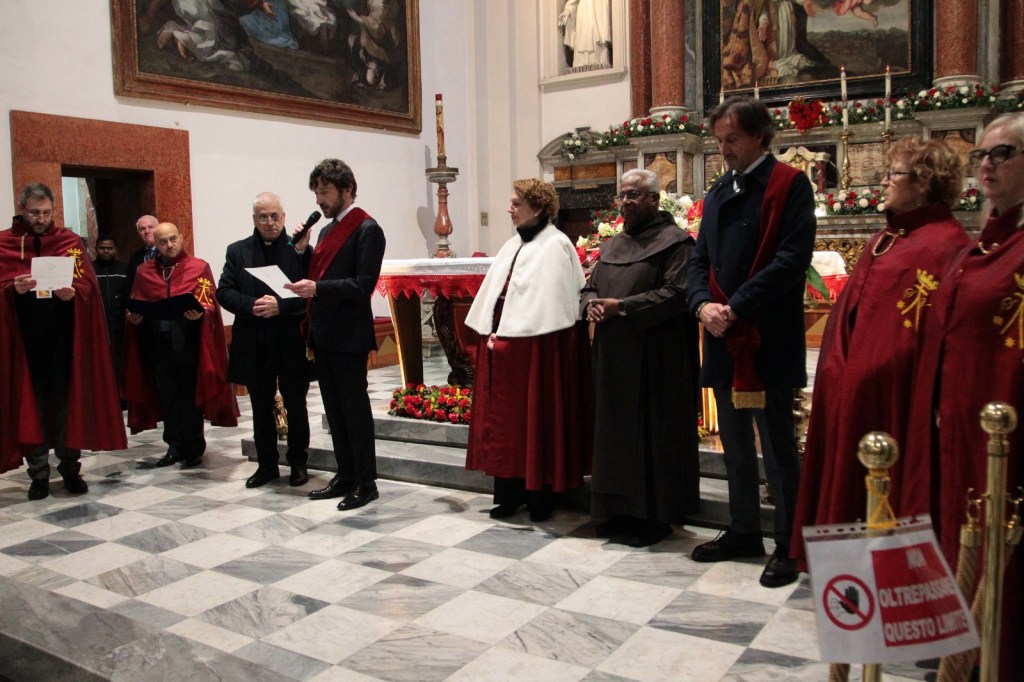 A group of individuals in formal attire, many wearing red cloaks, stand in a church setting. A man at the center speaks into a microphone while others listen attentively. The background features religious artwork and floral decorations.