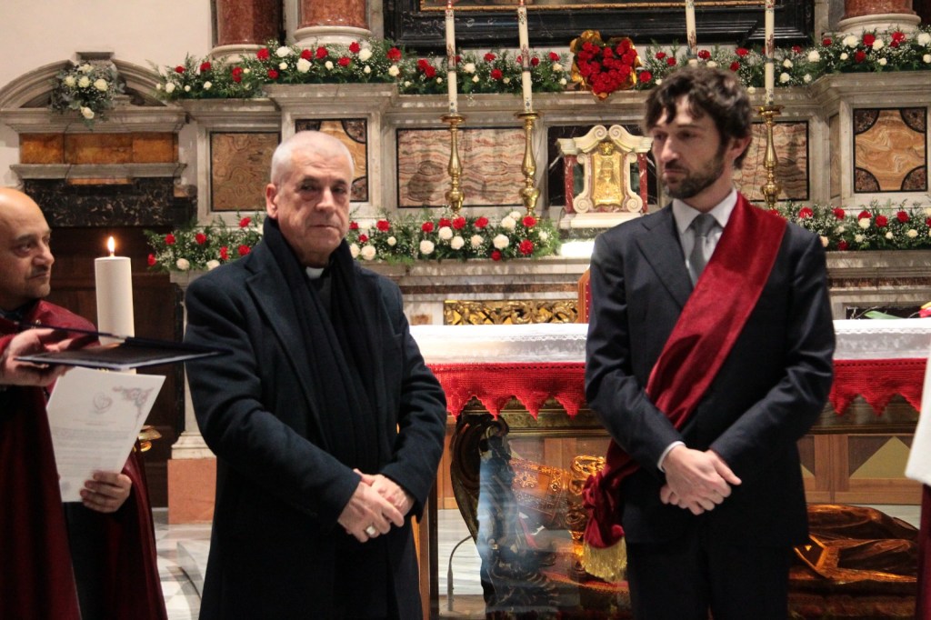 Two men stand in a church setting during a ceremony, with a third person partially visible on the left holding a document. The background features floral decorations and a candle.
