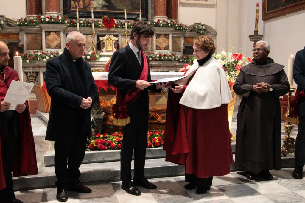 A formal ceremony taking place in a church, featuring several individuals in traditional attire. One person is presenting a document to another, while others look on. The background includes floral arrangements and religious decor.