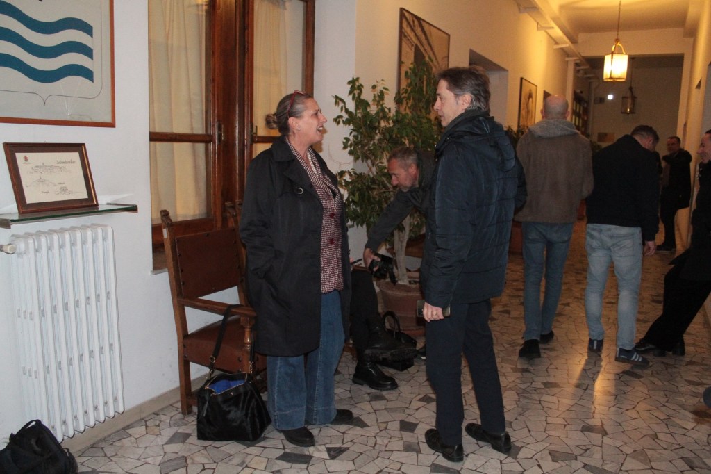 A group of people interacting in a hallway, with one woman wearing a black coat and talking, while others are engaged in conversation and activities in the background. The setting features tiled flooring, framed documents on the wall, and a radiator.