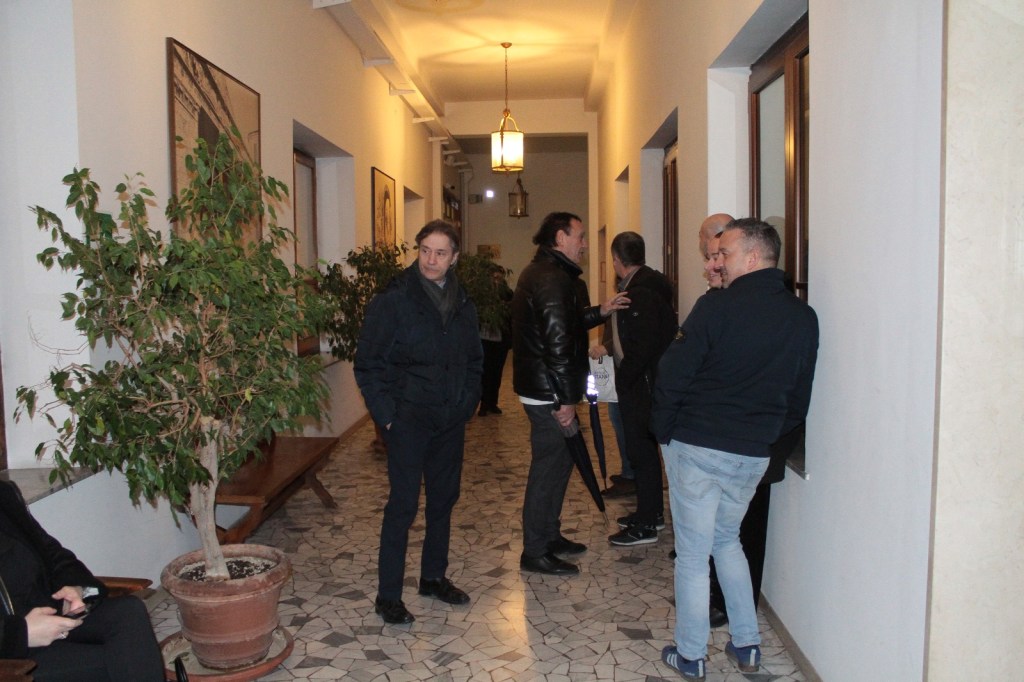 A group of men chatting in a hallway with potted plants and decorative artwork on the walls.
