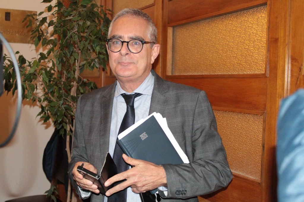 A man in a gray suit holding a notebook and a smartphone, standing in a room with wooden walls and a leafy plant in the background.