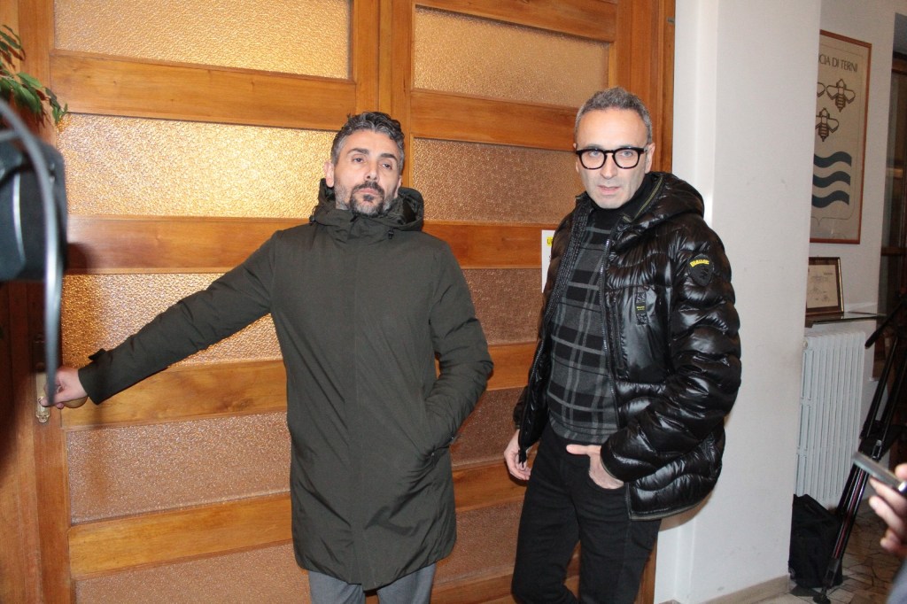 Two men standing near a wooden door, one wearing a dark coat and the other in a puffer jacket, both looking slightly serious.