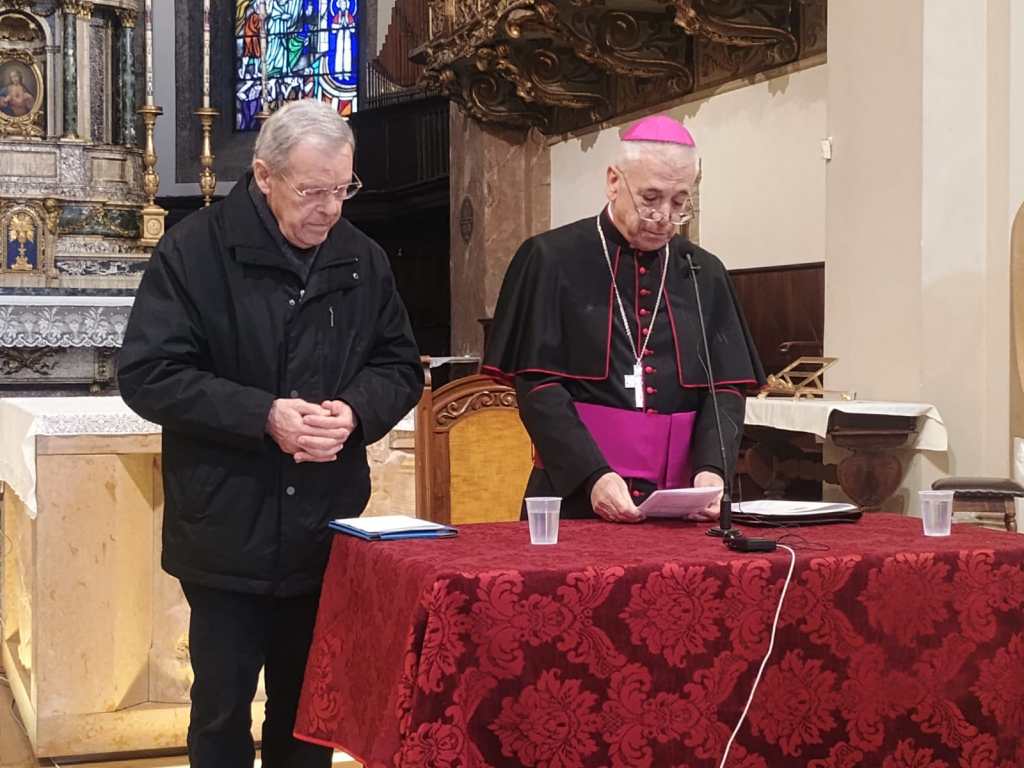 Two men standing at a table inside a church. One man in a black jacket stands to the left, while the other, wearing clerical attire with a pink cap, reads from a paper at the table.