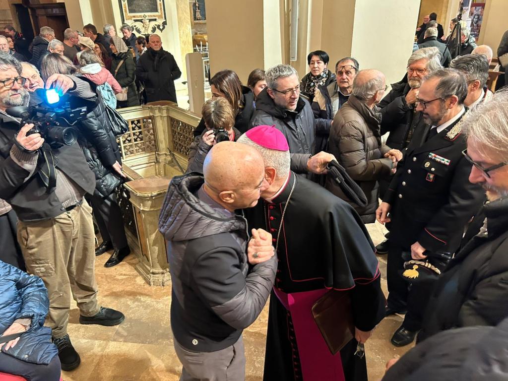A crowd of people is gathered in a church, with one individual embracing and greeting a religious leader, who is wearing ceremonial attire. Photographers capture the moment as attendees look on.