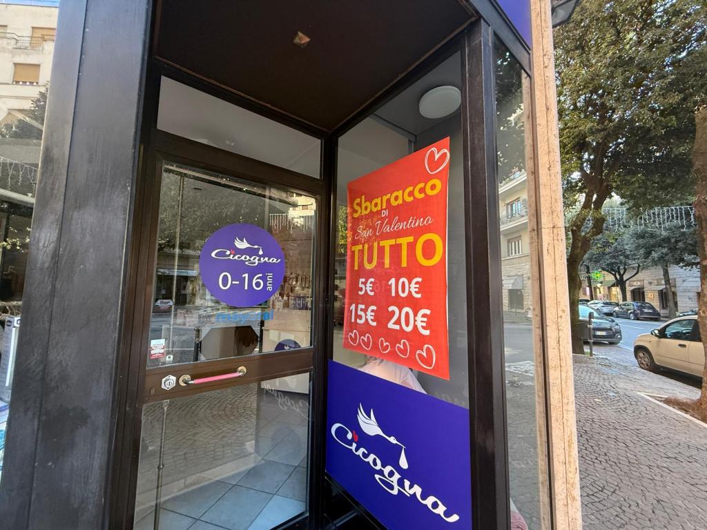 Exterior view of a store entrance with promotional signs for a Valentine's sale, featuring prices ranging from 5€ to 20€. The store is named 'Cicogna' and targets children aged 0-16 years.