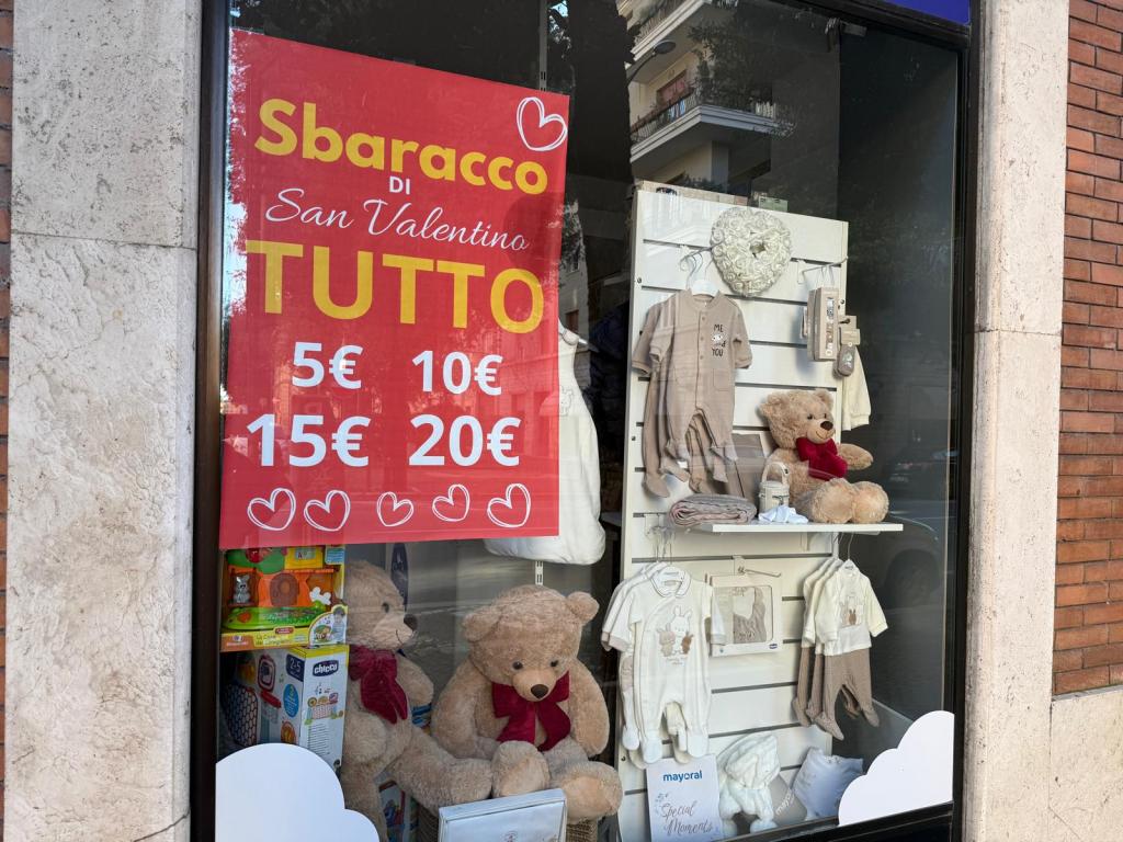 Store window display promoting a Valentine's Day sale with various children's clothing and teddy bears, featuring prices of 5€, 10€, 15€, and 20€.