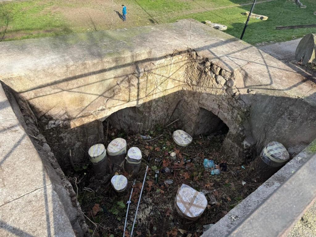 An above-ground view of a sunken area showing circular concrete bases and debris, with a person walking in the background.
