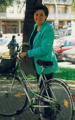 A woman wearing a turquoise blazer is smiling while standing next to a bicycle in an outdoor setting.