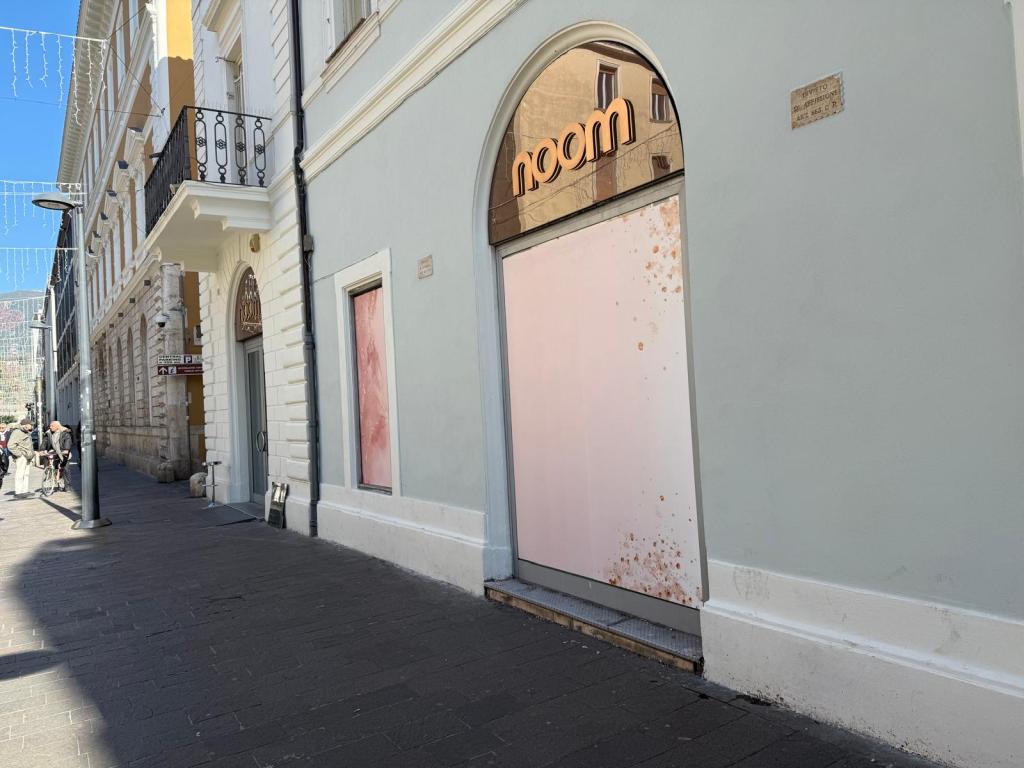 A storefront featuring the word 'noom' above a large, partially covered pink door, surrounded by light-colored buildings on a pedestrian street with a few people walking by.