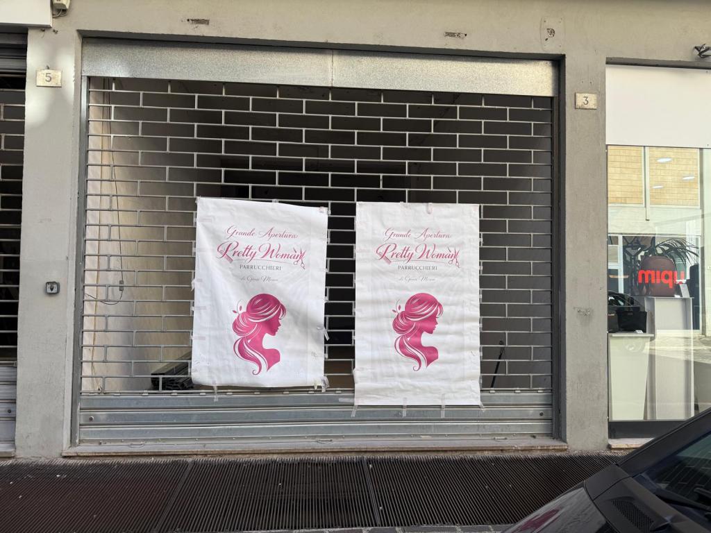 Exterior view of a salon called 'Pretty Woman' featuring two banners with a pink hair silhouette design, positioned in front of a closed shop with a metal grate.