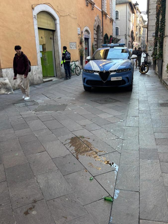 A blue car parked in a narrow street with a pedestrian walking by, a man in uniform nearby, and a spilled liquid on the cobblestone pavement.