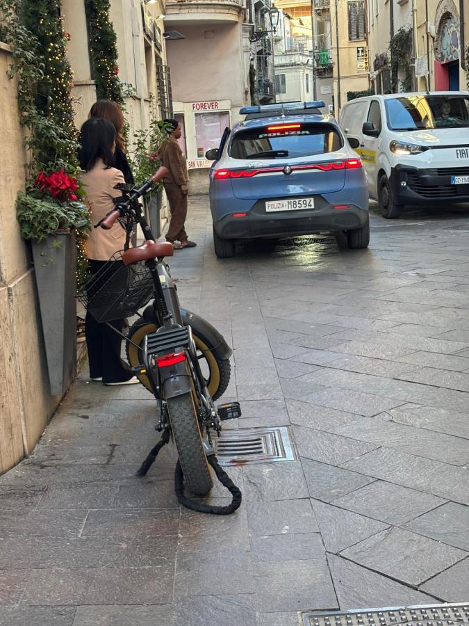 A narrow street scene featuring a parked bicycle on the left, with two women standing nearby. In the background, a blue vehicle with a police insignia is backing up next to a white van.