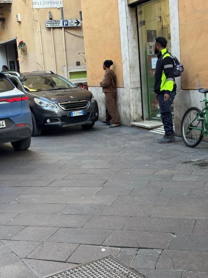 A street scene featuring two individuals near a parked car, one standing and the other leaning against a wall, with a bicycle nearby and a sign indicating a parking direction.