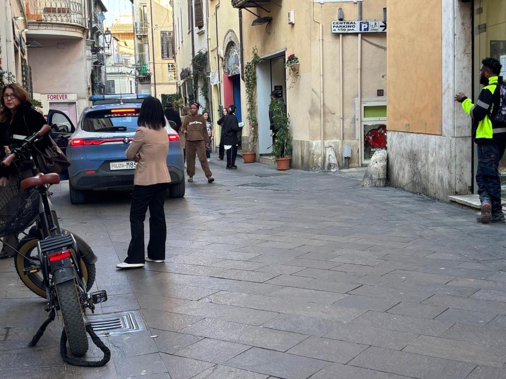 A narrow street scene featuring people interacting and a police car parked. An electric bicycle is shown in the foreground, and shops line the background walls.