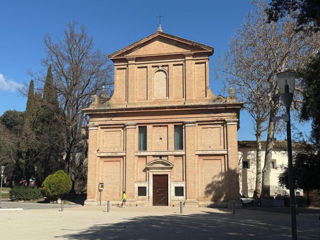 A red-brick building with a triangular roof and a small cross at the top, set against a clear blue sky. The structure is surrounded by trees and a paved area, and a person is visible walking in front of the building.