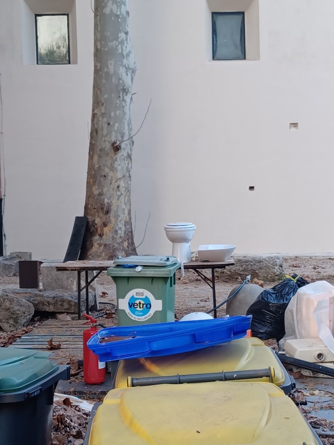 Scene of a cluttered outdoor area featuring various trash bins, a toilet, and other discarded items near a tree and a wall.