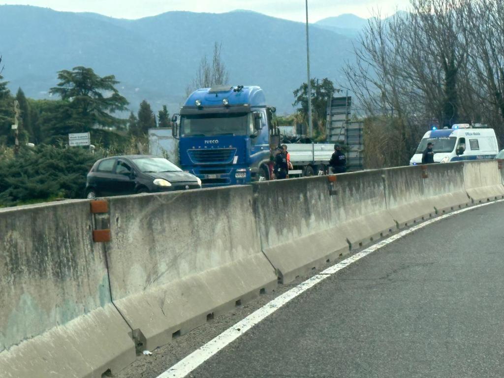 A blue truck and a black car are stopped on the side of a road near a barrier, with police and emergency vehicles in attendance.