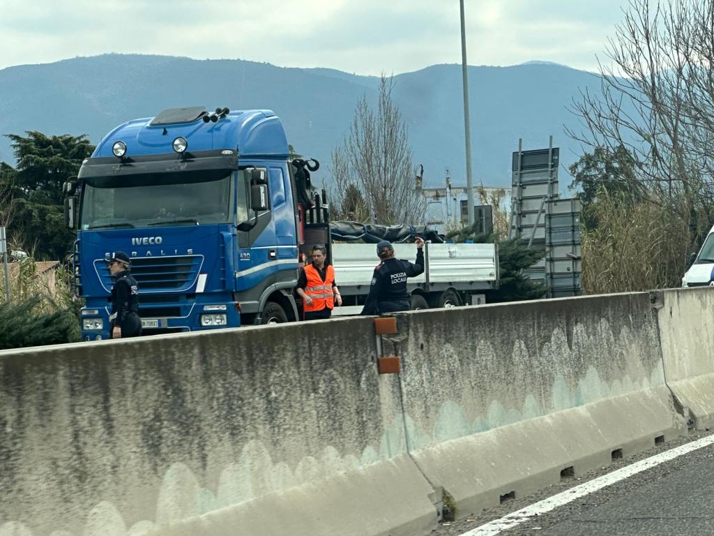 A blue Iveco truck parked along the highway, with two individuals in safety vests interacting near a white truck, under a cloudy sky and mountains in the background.