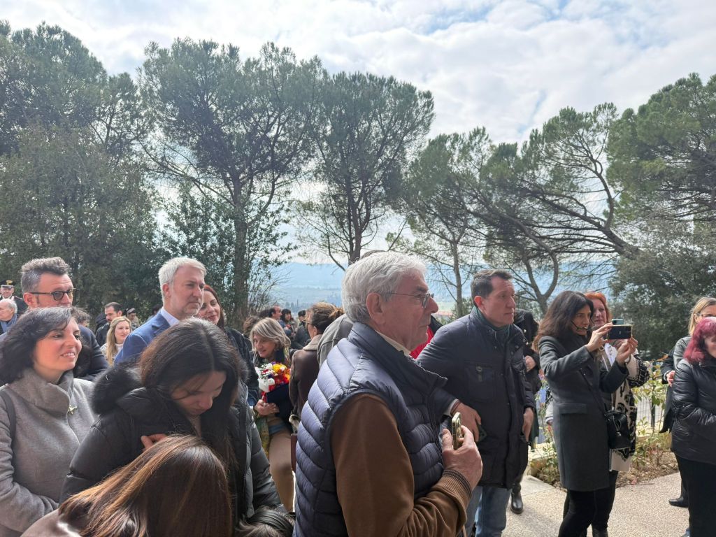 A crowd of people gathered outdoors, with some taking photos and others engaged in conversation, surrounded by trees and a cloudy sky.