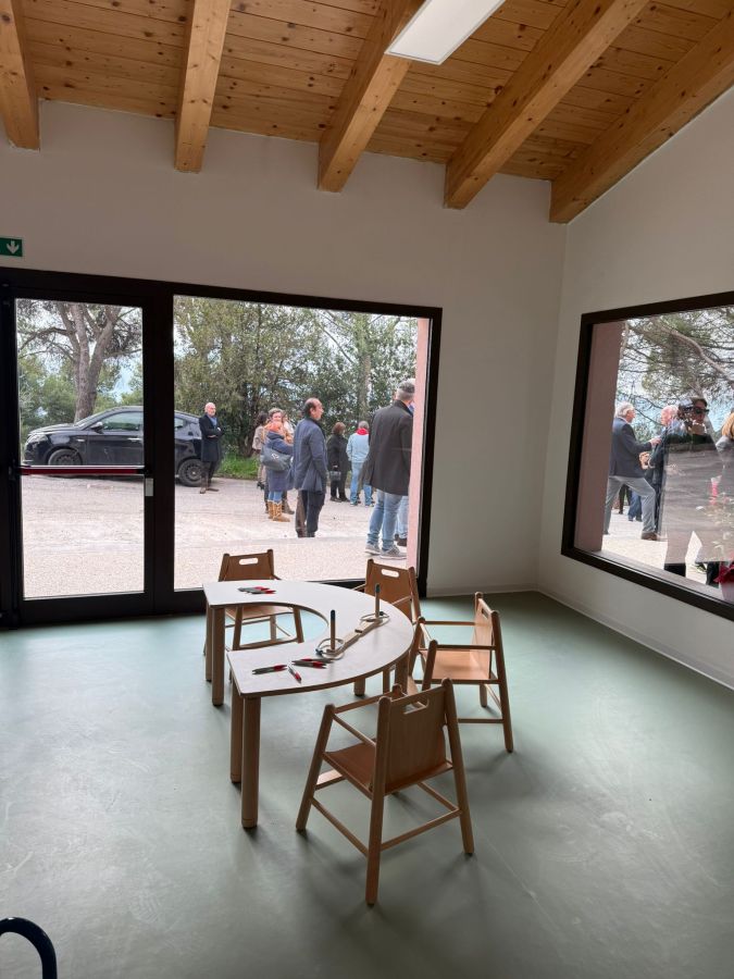 Interior view of a room with wooden beams, featuring a table and chairs for children. Outside the windows, a group of people is seen conversing.