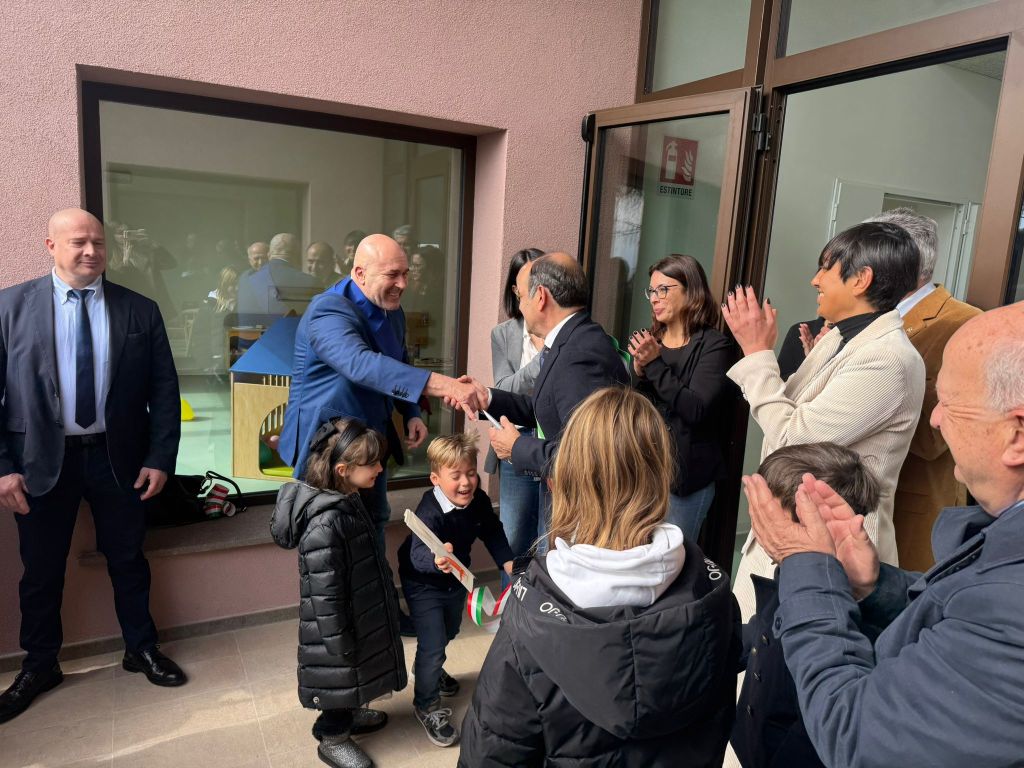 A group of people celebrating outside a building, with two men shaking hands while surrounded by children and adults applauding.