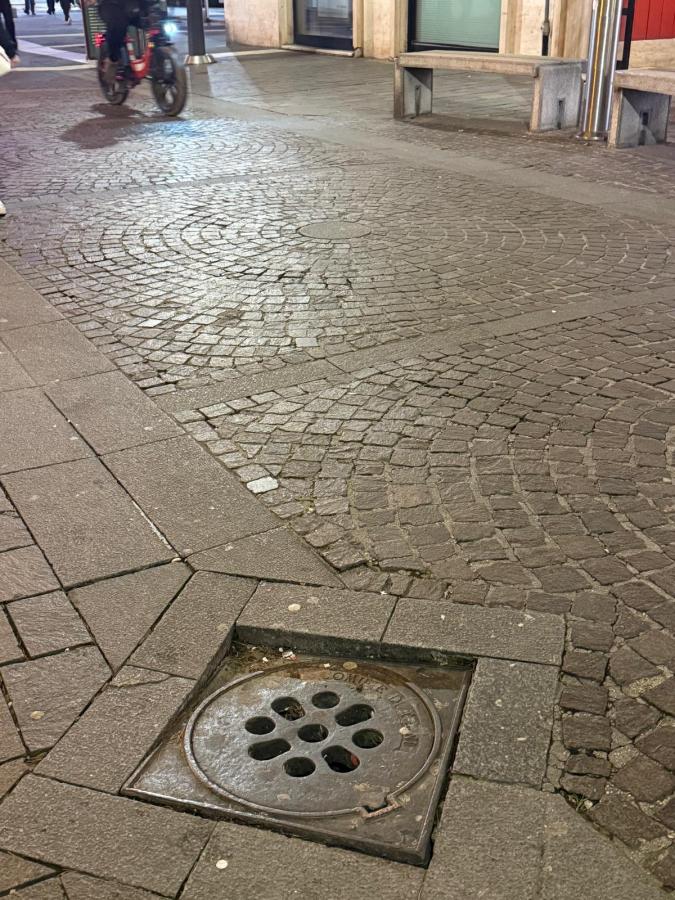 A cobblestone street at night with a visible manhole cover surrounded by patterned stone pavement and a cyclist in the background.