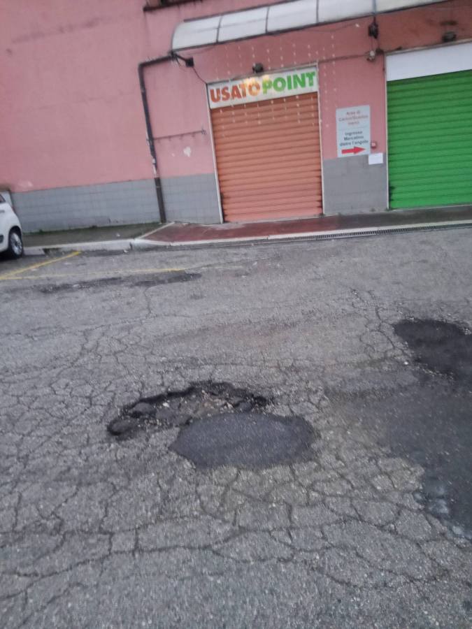 Exterior view of a building with a sign reading 'USATO POINT' above an orange shutter door, with a green shutter door to the right and a parking area with visible potholes and cracked pavement.