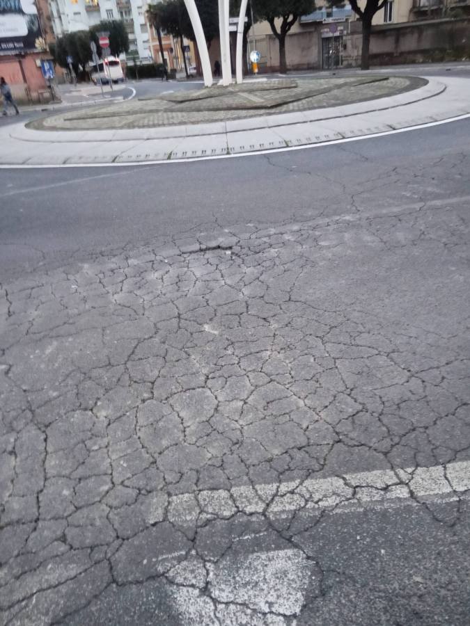 A cracked asphalt surface of a road leading into a circular roundabout with trees and a decorative center structure.