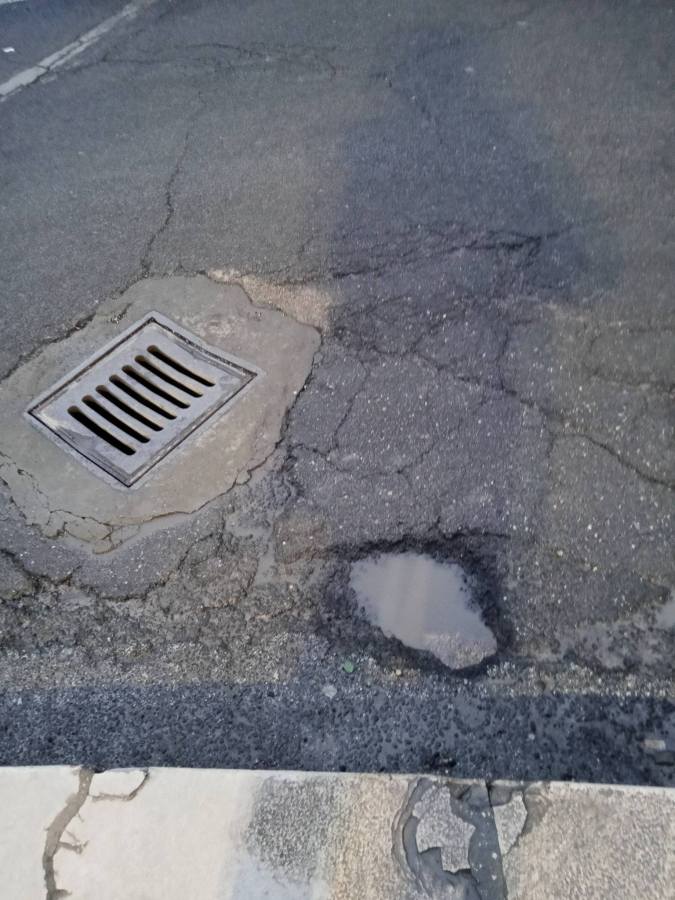 A view of a cracked pavement with a metal drainage grate on the left and a small puddle on the right.