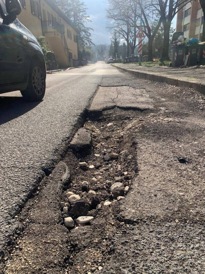 A close-up view of a damaged road showing a large pothole filled with stones and gravel, alongside a street with parked cars and residential buildings.
