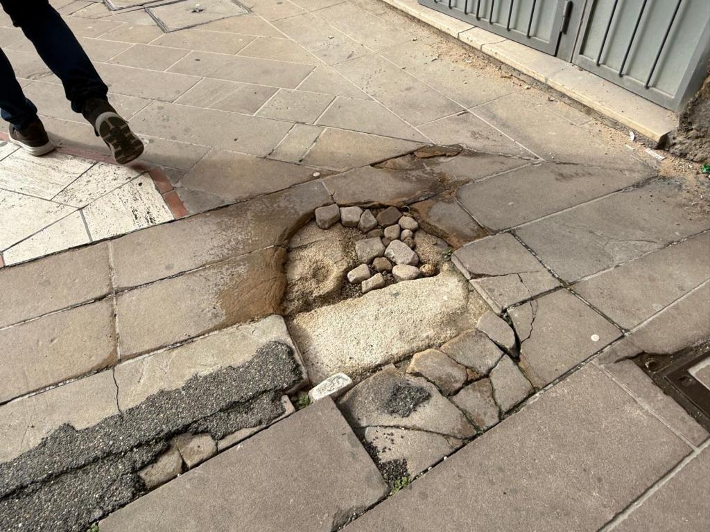 A sidewalk showing a large, uneven pothole filled with rocks and water, with a person walking nearby.