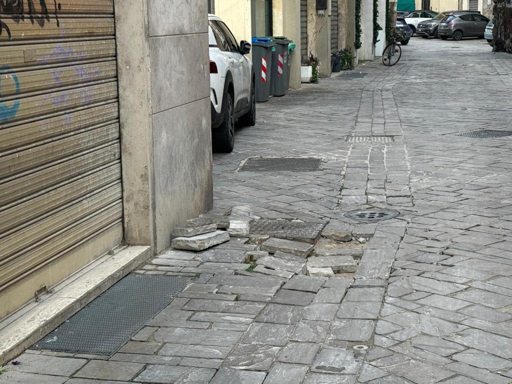 A cobblestone alleyway showing uneven pavement with raised stone slabs, a parked white car to the right, and a bicycle nearby.