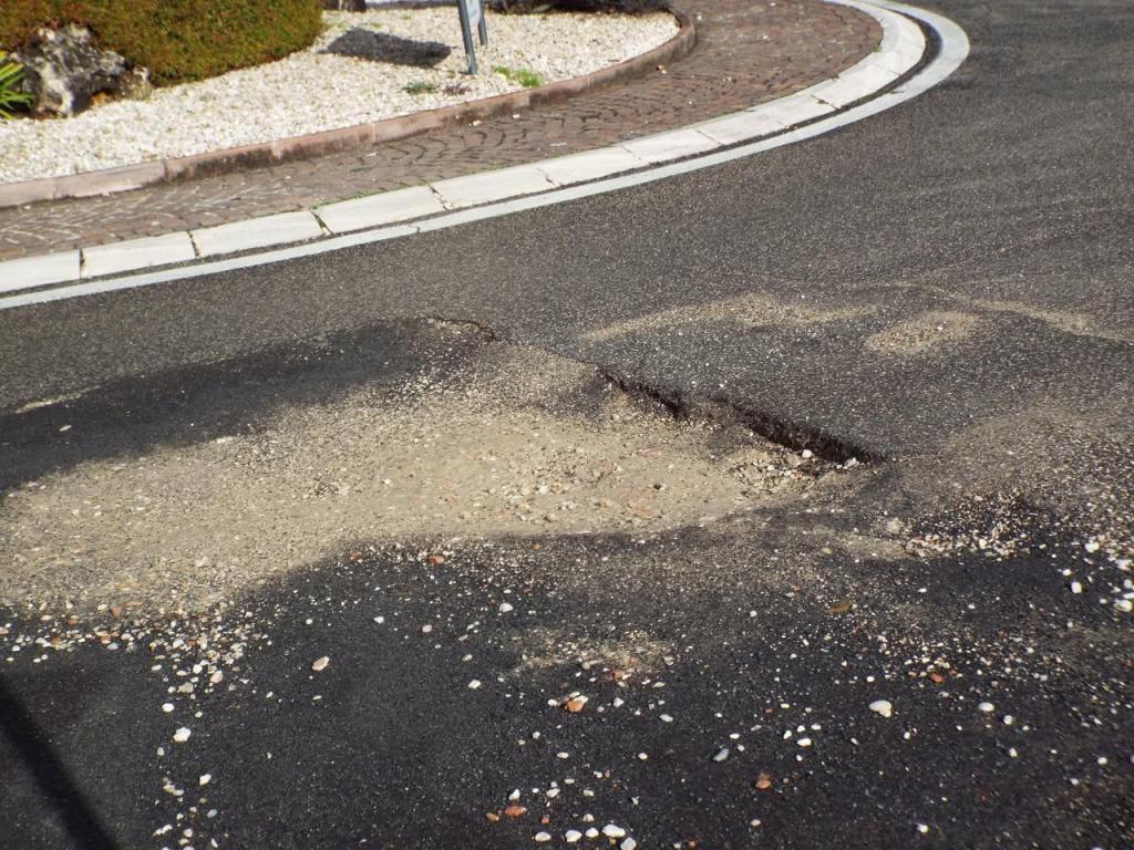 A damaged section of asphalt road showing a pothole filled with loose gravel and dirt.