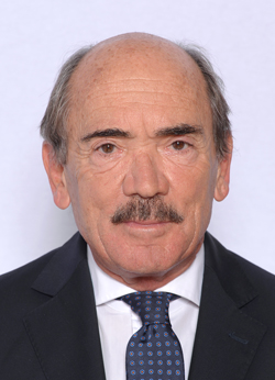 Headshot of a man with short hair and a mustache, wearing a dark suit and a patterned tie, against a plain background.
