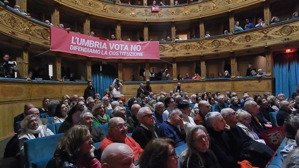 A crowded theater filled with people listening attentively, with a prominent pink banner that reads 'L'UMBRIA VOTA NO DIFENDIAMO LA COSTITUZIONE' in the background.