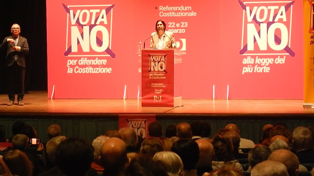 A public speaking event where a woman stands at a podium labeled 'VOTA NO,' addressing an audience while a man interprets in sign language beside her. The backdrop features bold red and white graphics promoting a constitutional referendum.