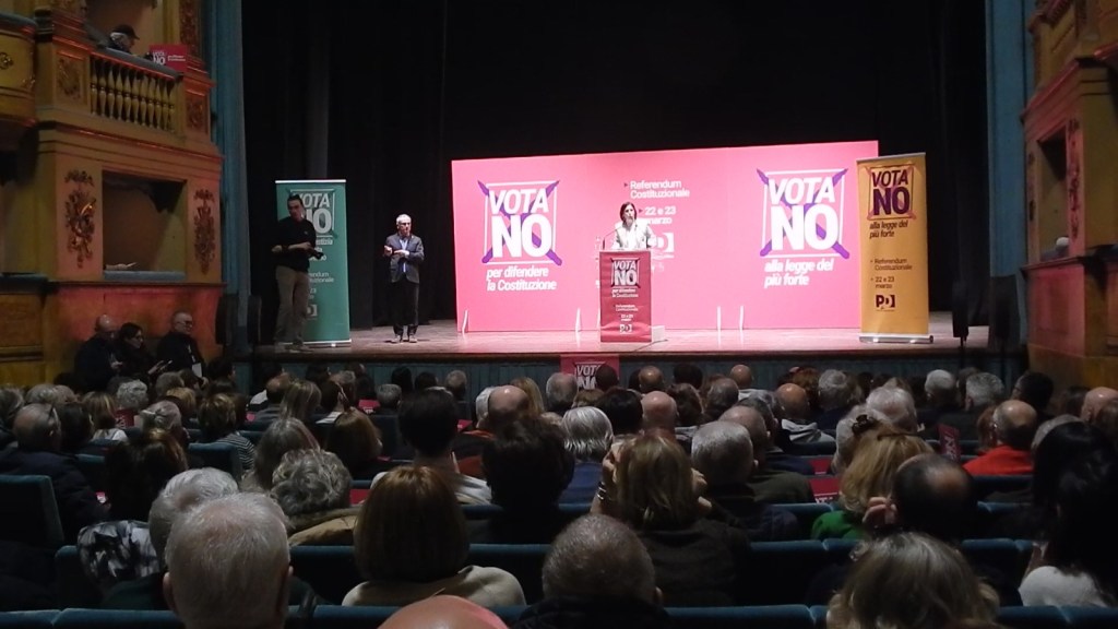 A political rally taking place in a theater, with a speaker addressing a seated audience. The backdrop features a large 'VOTA NO' sign related to a constitutional referendum, and there are banners around the stage promoting the 'NO' vote.